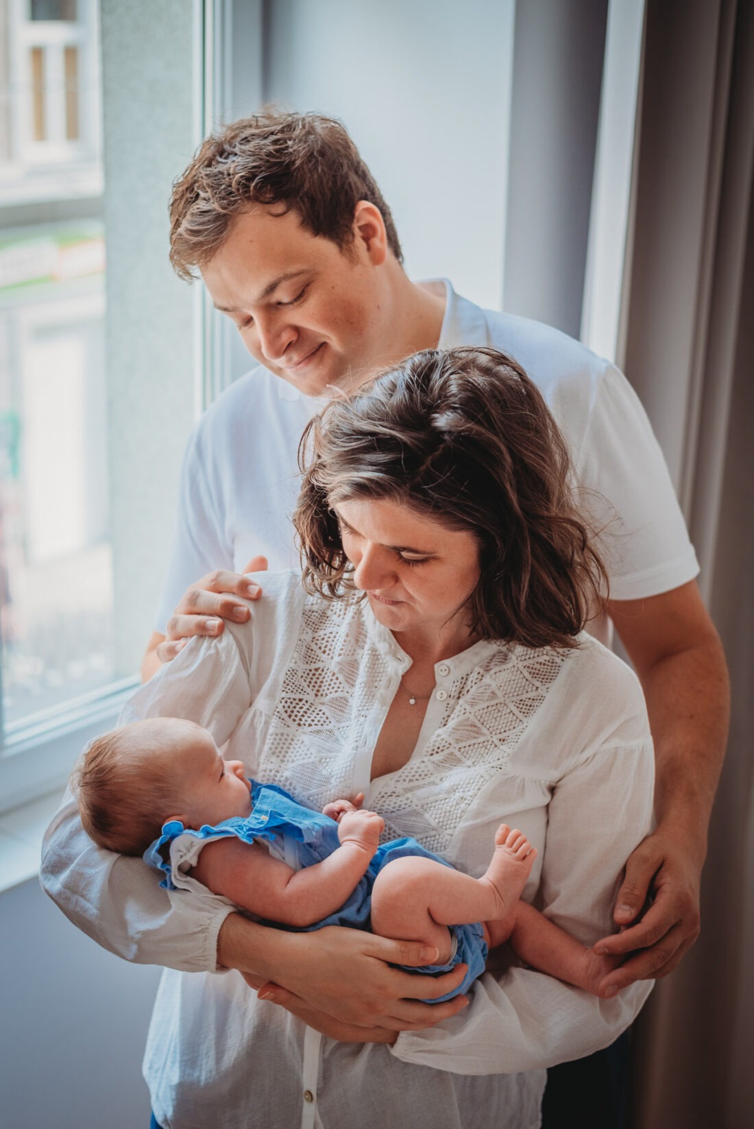 Parents standing by window holding newborn baby in natural light.