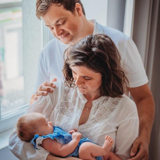 Parents standing by window holding newborn baby in natural light.