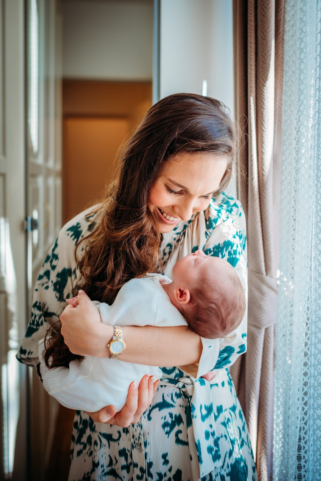 Smiling mother cradling her newborn by a sunlit window.