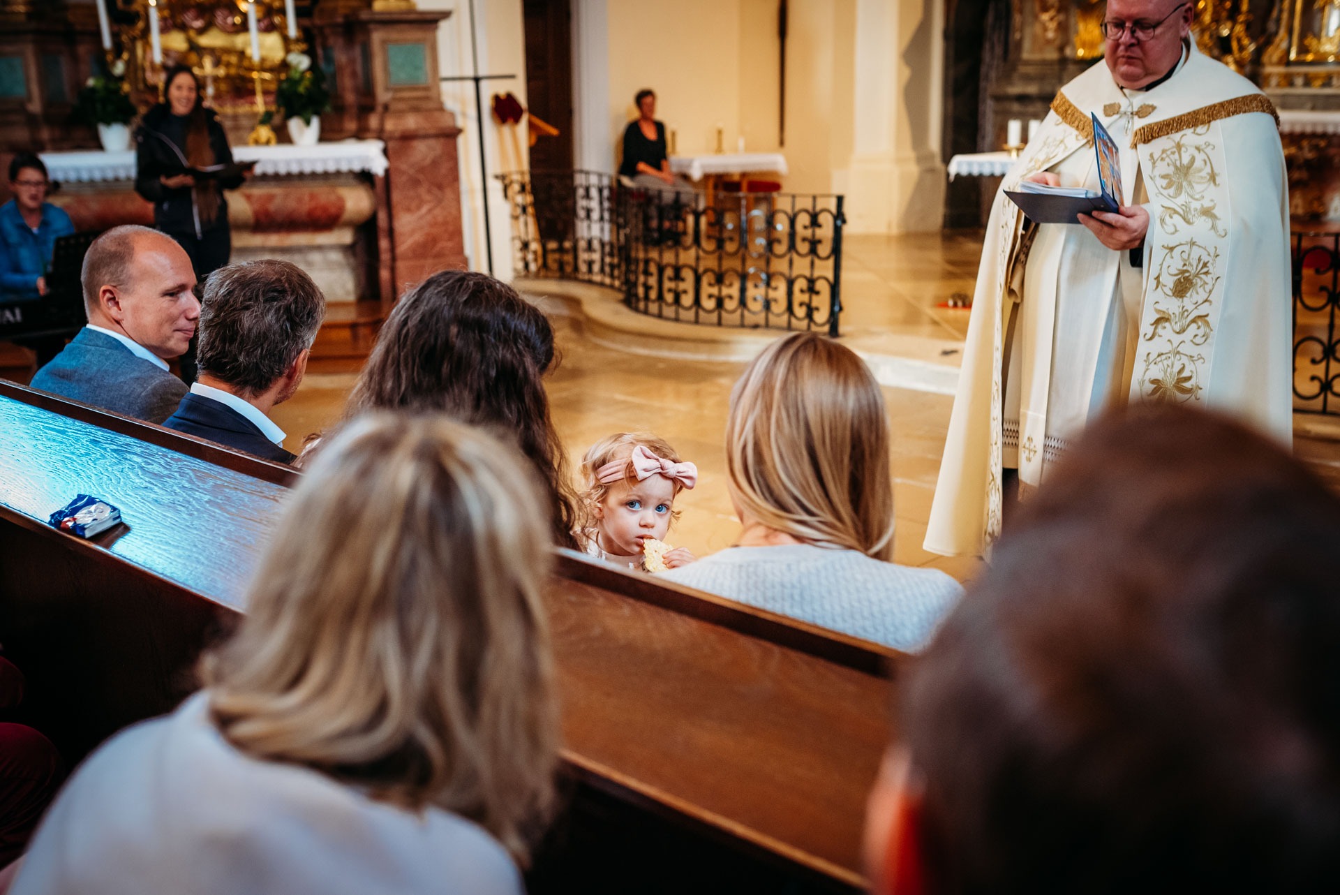 Toddler girl with pink bow looks back during church baptism captured by family event photographer