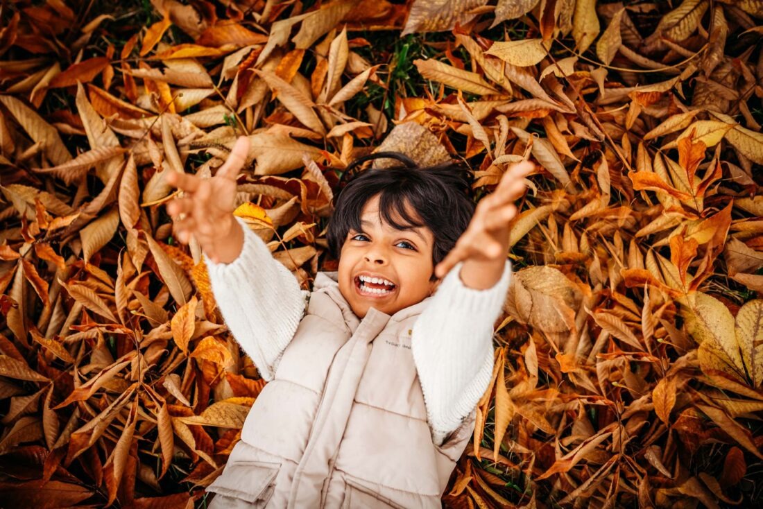 Young boy lying on the ground amidst autumn leaves, grinning with arms outstretched during a photo shoot in Prague.
