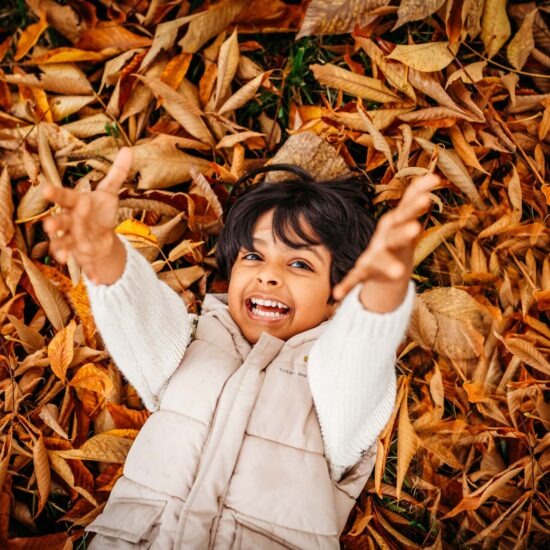 Young boy lying on the ground amidst autumn leaves, grinning with arms outstretched during a photo shoot in Prague.