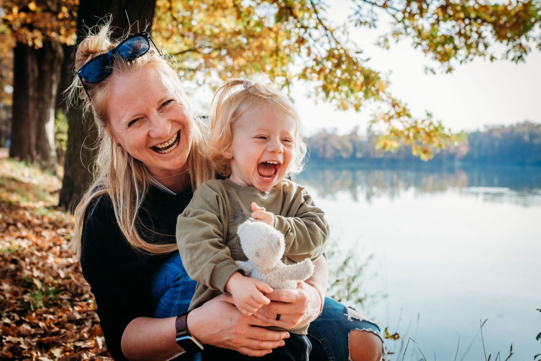 A mother and her young daughter share a joyful laugh, cuddling together under the autumn trees with golden leaves in the background.