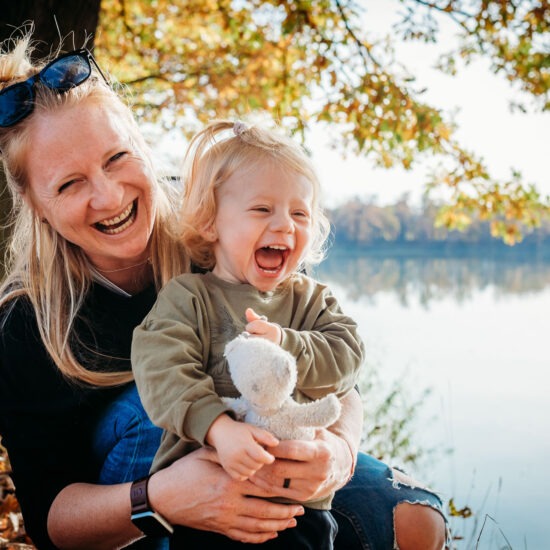 A mother and her young daughter share a joyful laugh, cuddling together under the autumn trees with golden leaves in the background.