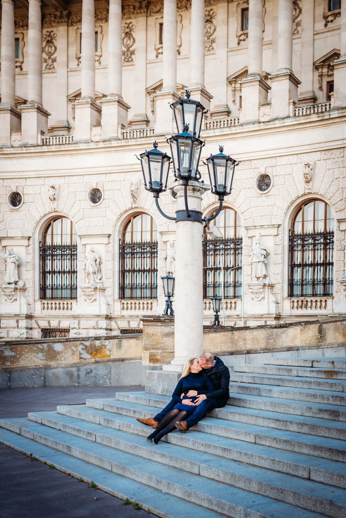 A pregnant couple sitting on the grand stairs of Hofburg Palace, enjoying their time together during their pregnancy photoshoot in Vienna.