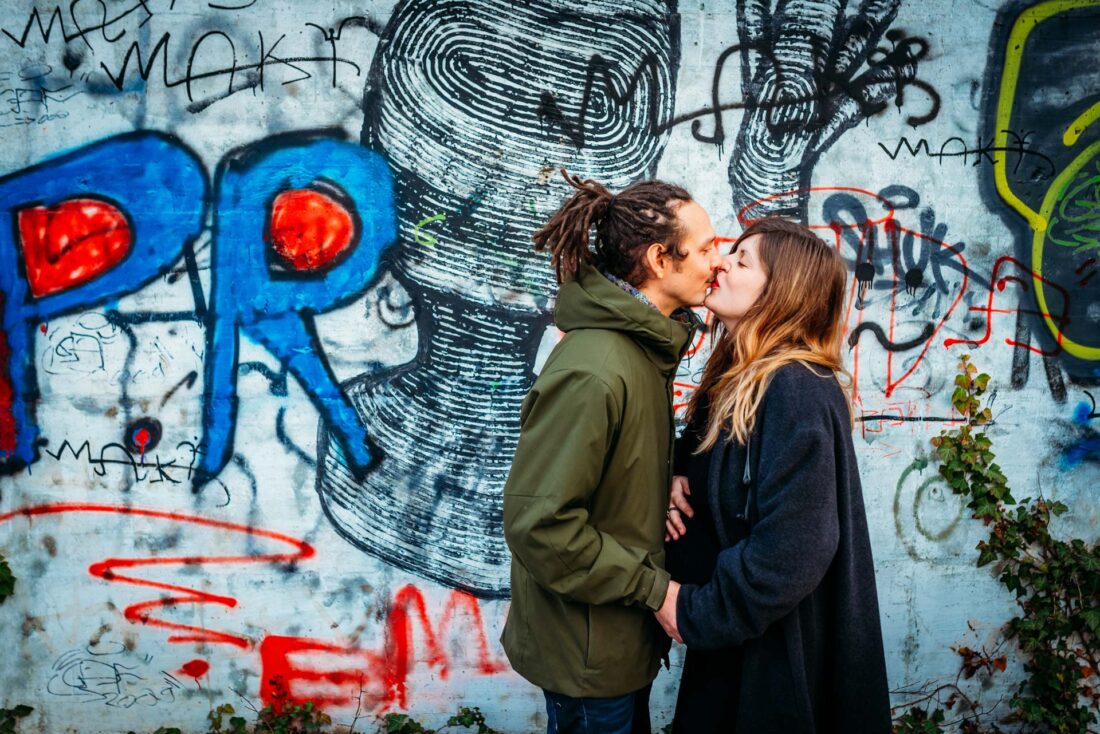 An expecting couple shares a kiss in front of a graffiti wall, blending maternity photography with an urban feel.