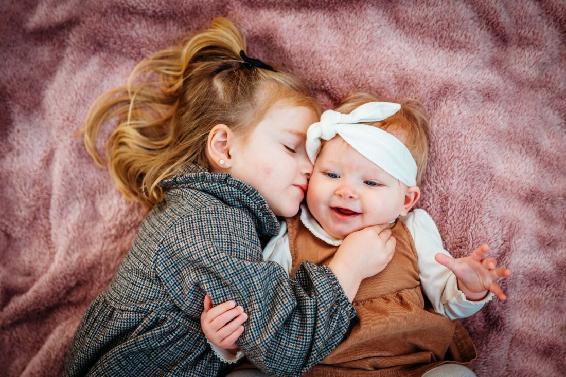 A touching close-up of a young girl gently cuddling her baby sister while lying on a soft, pink blanket outdoors.