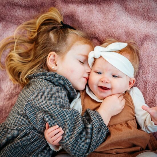 A touching close-up of a young girl gently cuddling her baby sister while lying on a soft, pink blanket outdoors.
