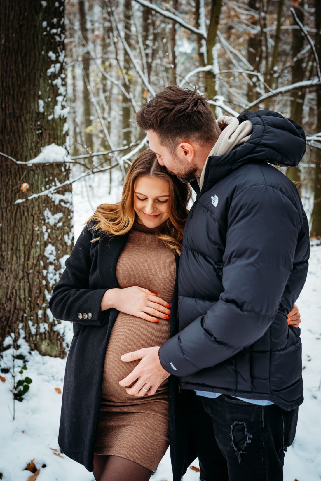 A father-to-be gently kisses his partner’s forehead as she smiles, enjoying a quiet moment in their snowy maternity photo shoot.