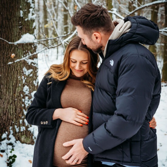 A father-to-be gently kisses his partner’s forehead as she smiles, enjoying a quiet moment in their snowy maternity photo shoot.