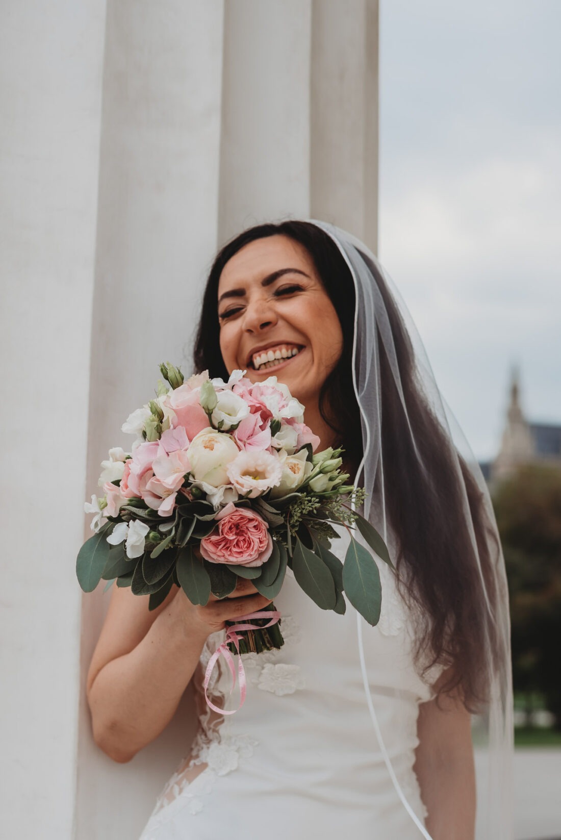 Bride laughing joyfully while holding her wedding bouquet close to her face, standing in front of a white column of the Theseus Temple in Vienna.