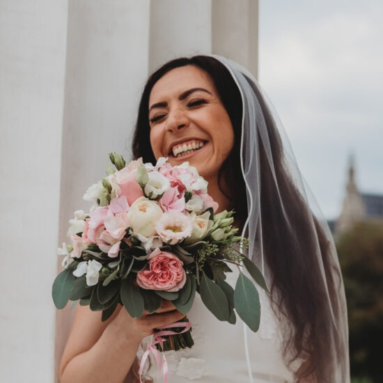 Bride laughing joyfully while holding her wedding bouquet close to her face, standing in front of a white column of the Theseus Temple in Vienna.