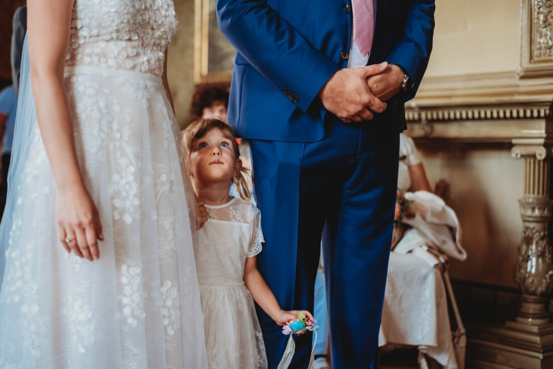 Bride and groom’s daughter holding her mother’s hand, looking up at her with curiosity as they stand together before the altar at their wedding in the Czech Republic.