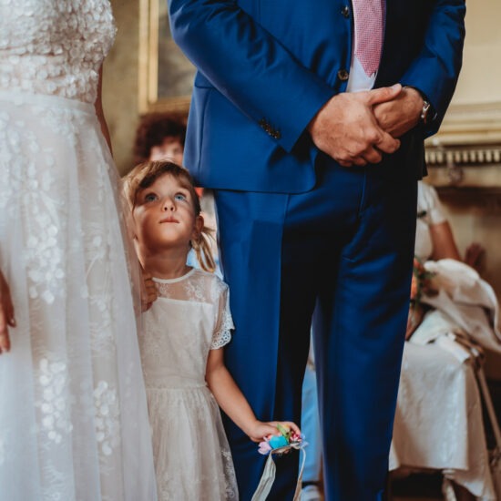 Bride and groom’s daughter holding her mother’s hand, looking up at her with curiosity as they stand together before the altar at their wedding in the Czech Republic.