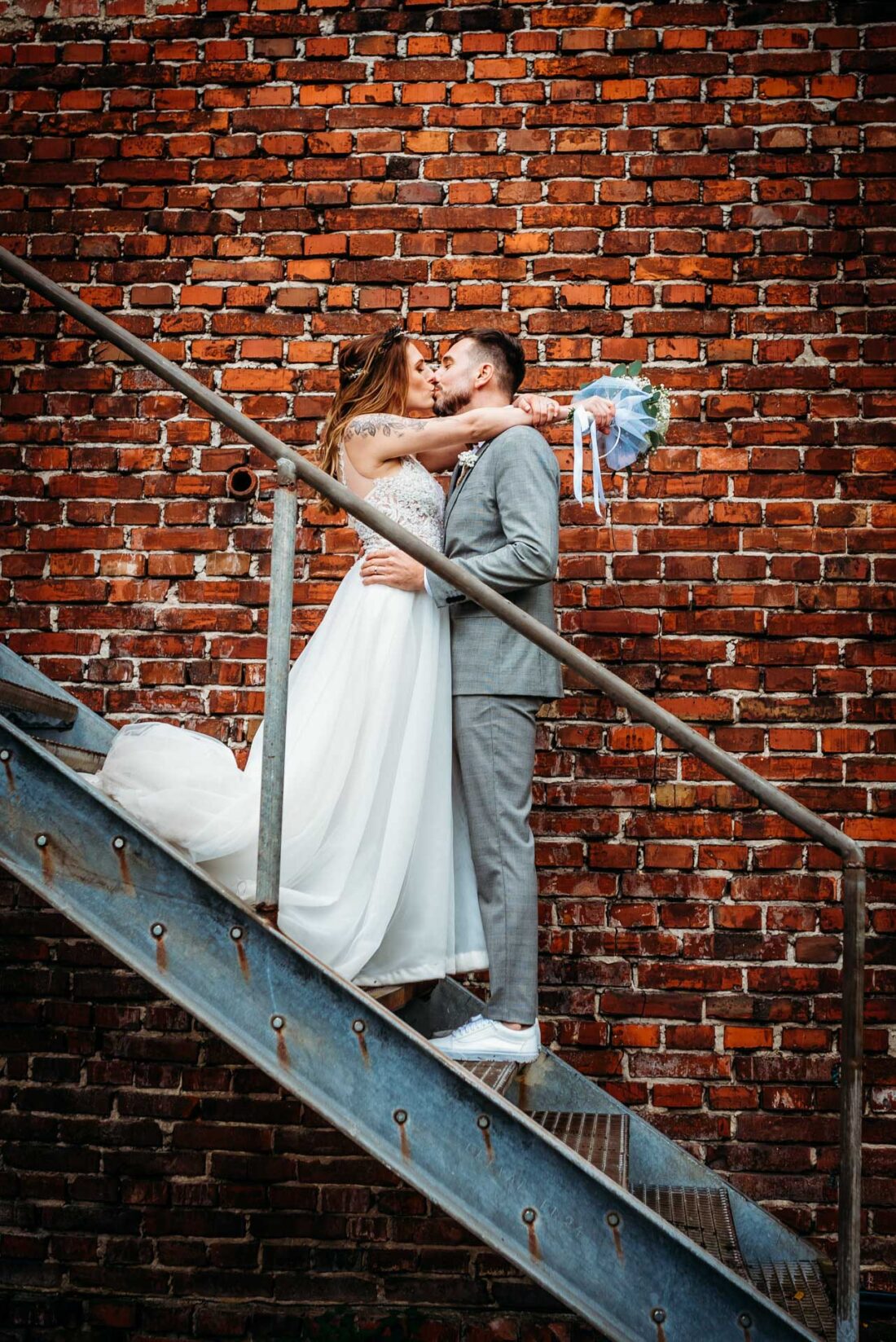 Newlyweds standing on metal stairs in front of a brick wall, with the bride resting her arms around her husband’s neck and kissing him.