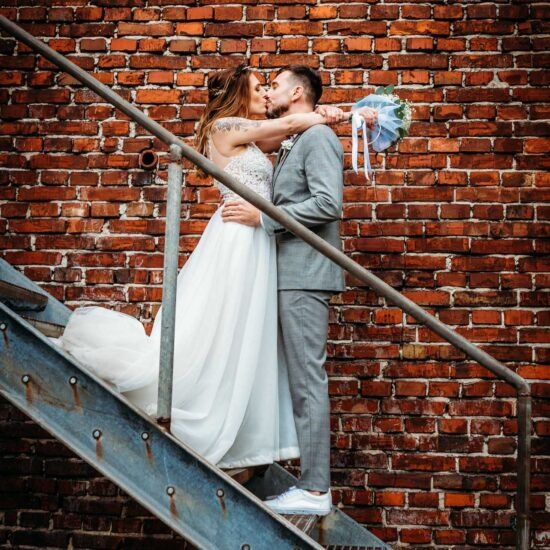 Newlyweds standing on metal stairs in front of a brick wall, with the bride resting her arms around her husband’s neck and kissing him.