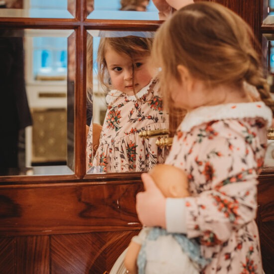 A young girl in a floral dress standing in front of a mirror, gazing at her reflection while holding her doll in one hand.