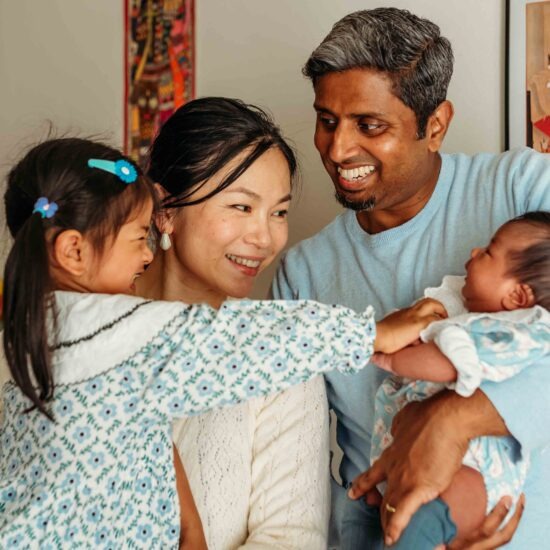 Young girl with her Chinese-Indian family, reaching out to touch her newborn sibling held gently by their father, captured by a family photographer in Vienna
