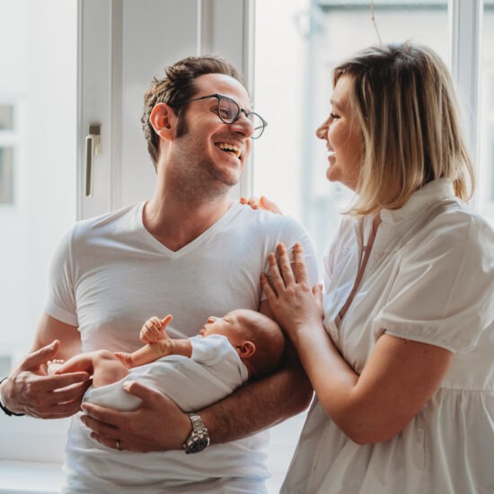 Smiling parents holding newborn baby by window during photo shoot in Vienna.