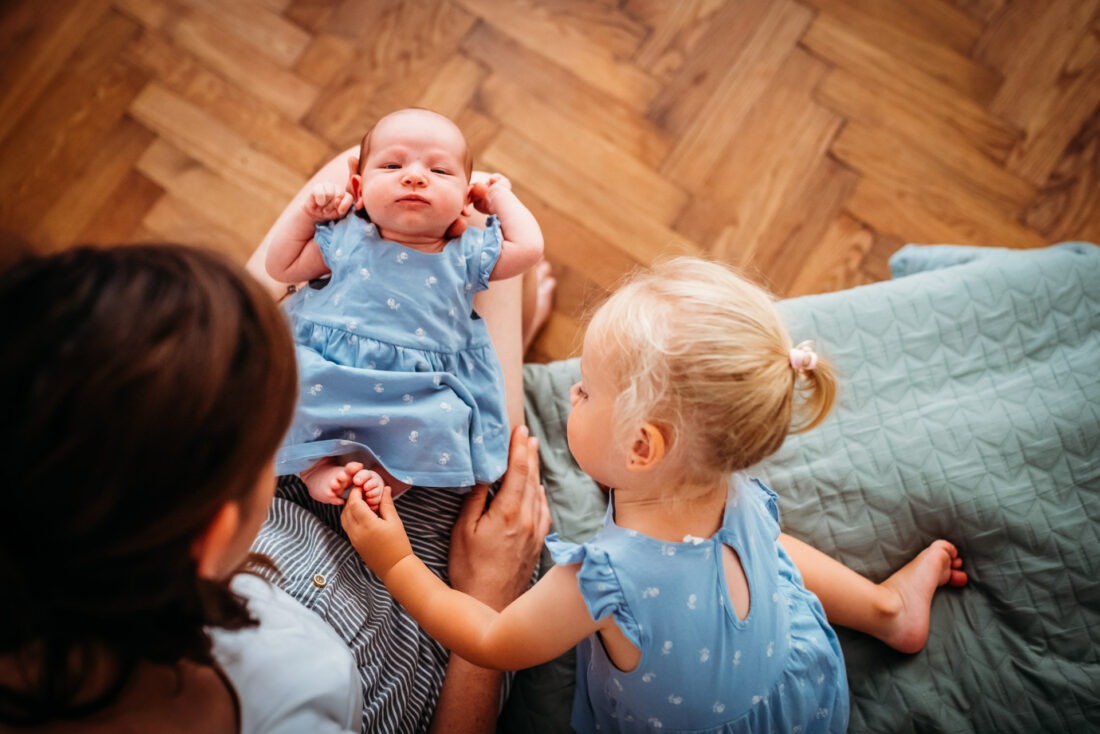 Toddler girl gently holding her baby sister’s foot during a newborn photo shoot at home.