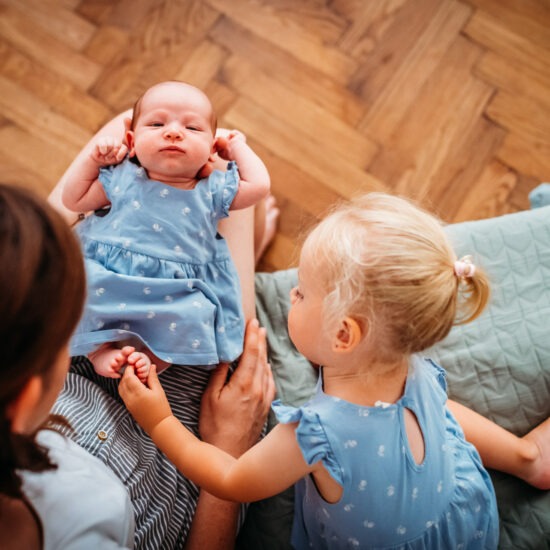 Toddler girl gently holding her baby sister’s foot during a newborn photo shoot at home.
