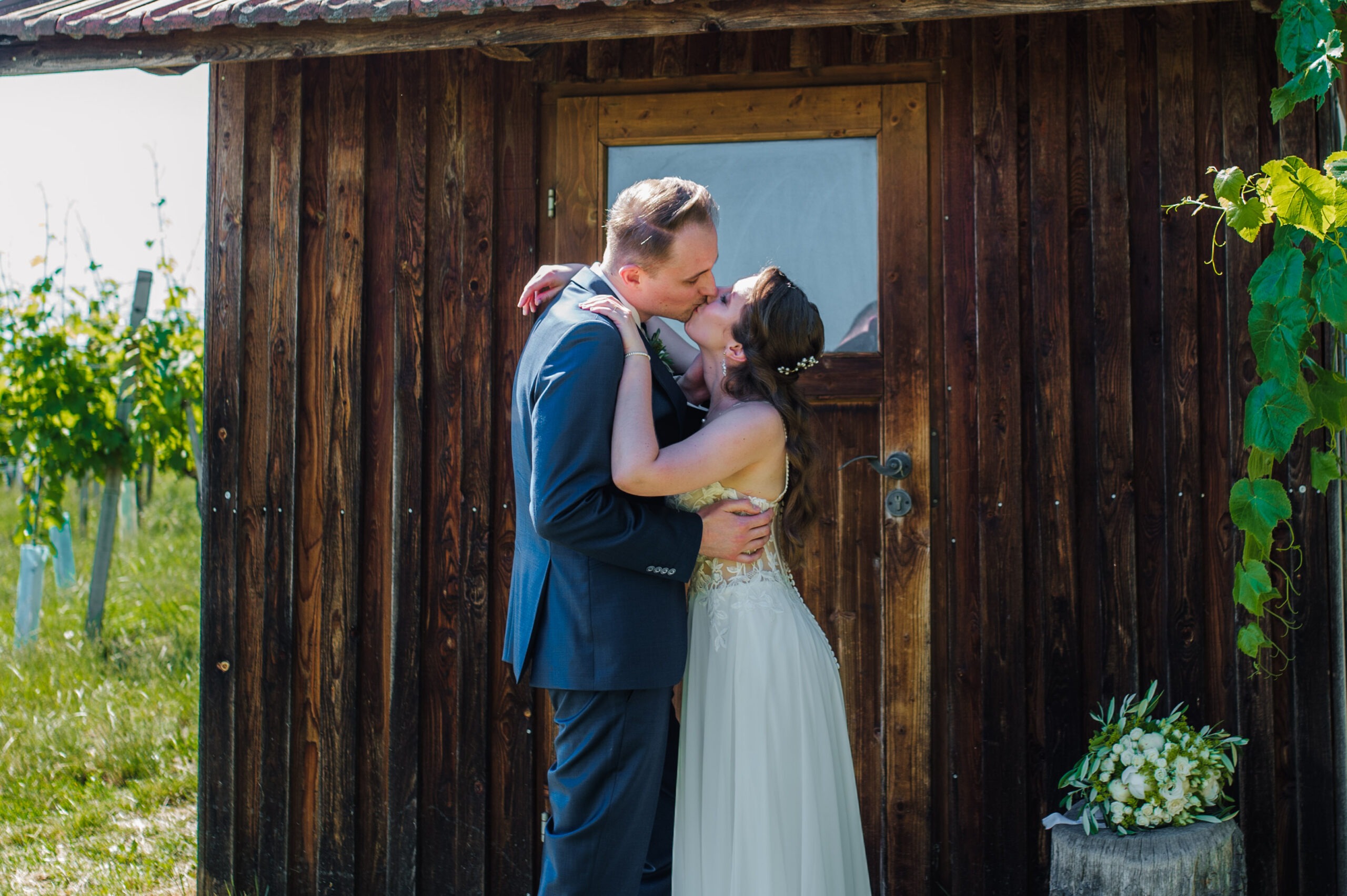 austria-vienna-summer-wedding-20 A newlywed couple embraces and kisses in front of a rustic wooden hut beside a vineyard, with the bride’s white-and-green bouquet resting nearby