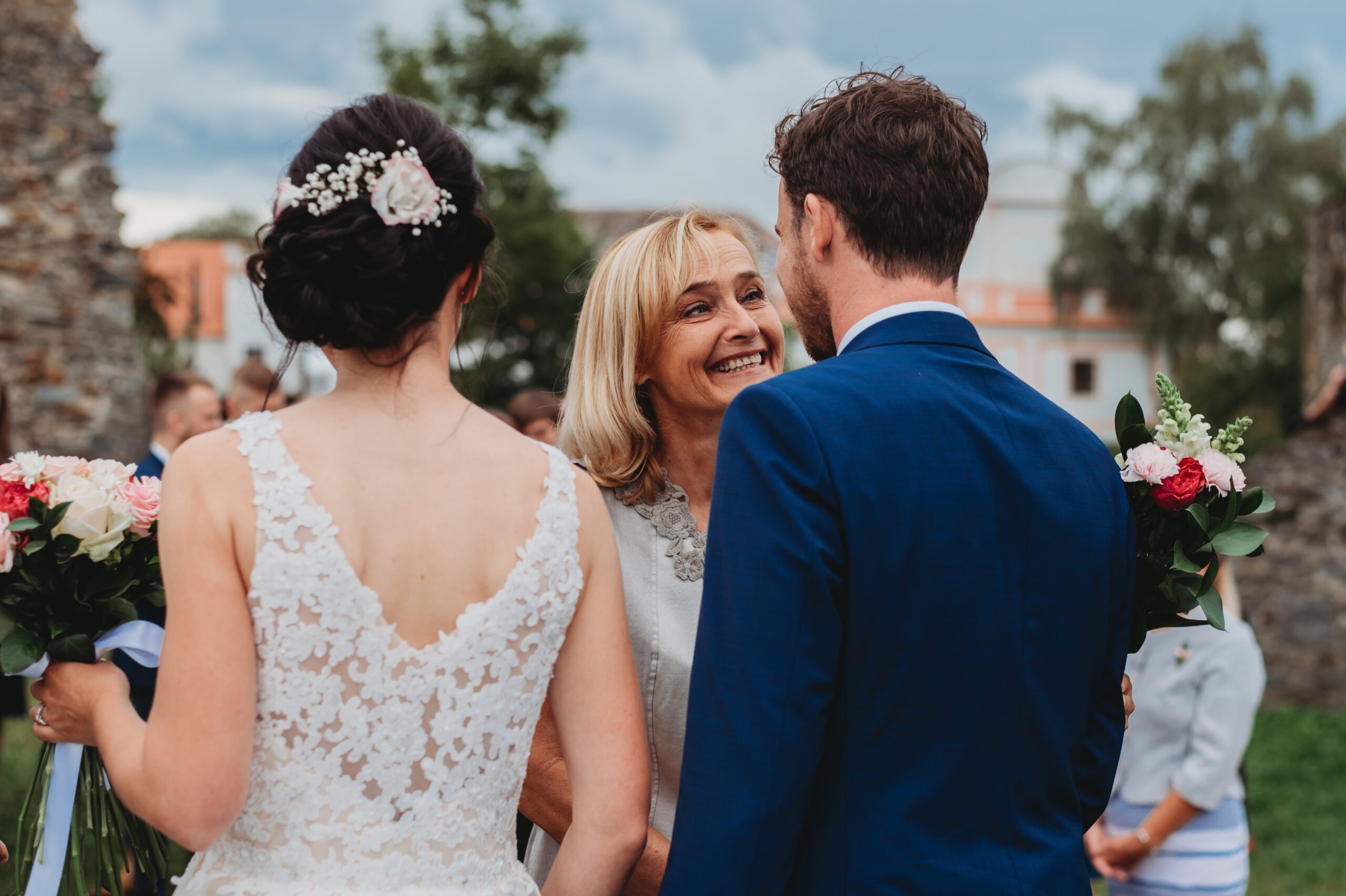 A smiling woman congratulates a newlywed couple after their wedding ceremony, standing between the bride in a lace dress and the groom in a blue suit.