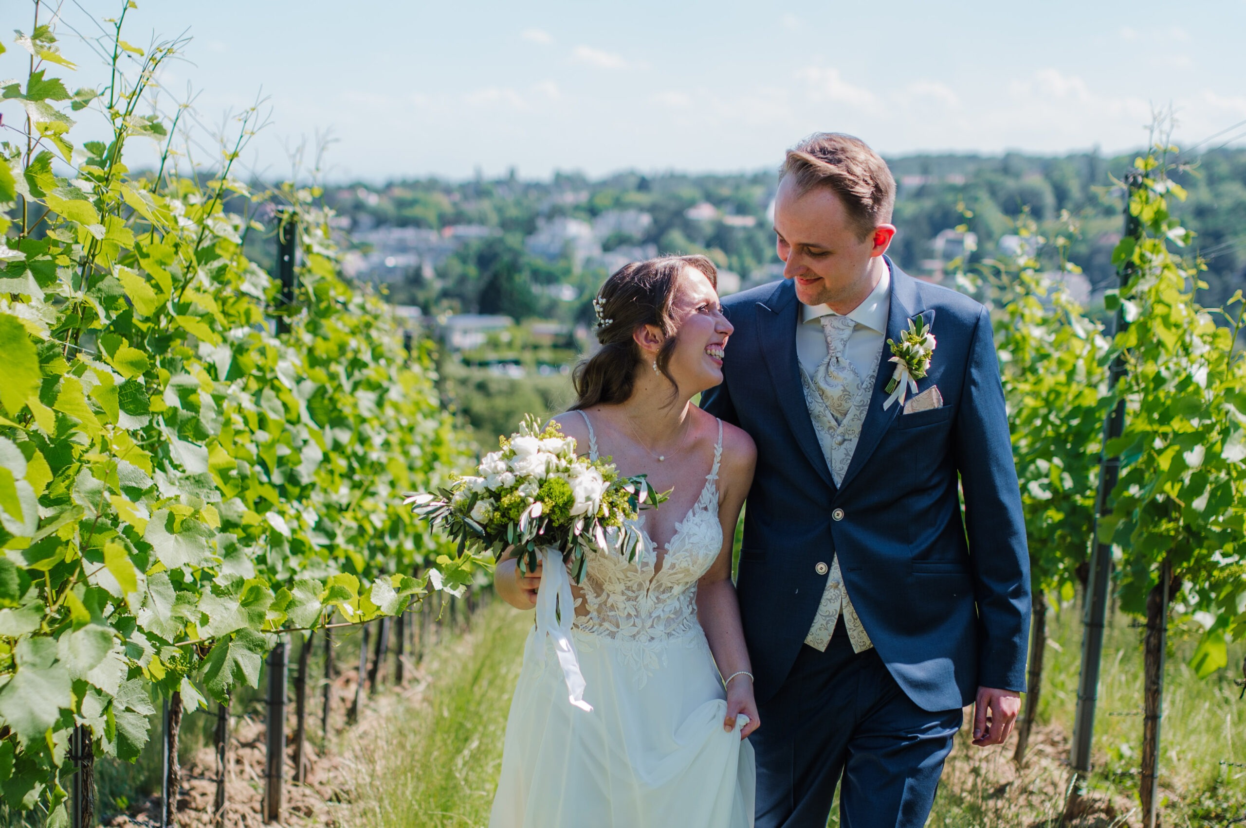 vienna-wedding-couple-first-look-vineyards A newlywed couple walks together through a sunlit vineyard, smiling at each other. The bride carries a white-and-green bouquet and wears a lace wedding dress, while the groom wears a navy suit